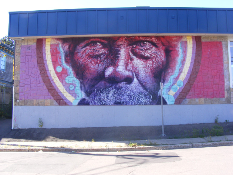The side of a building is painted with the face of an unhoused man. Only his eyes are visible as he stares at the viewer.