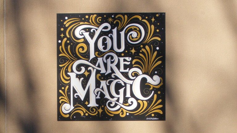 A square mural is positioned on a white wall. The mural is black with white lettering that reads "You Are Magic" with yellow swirls.