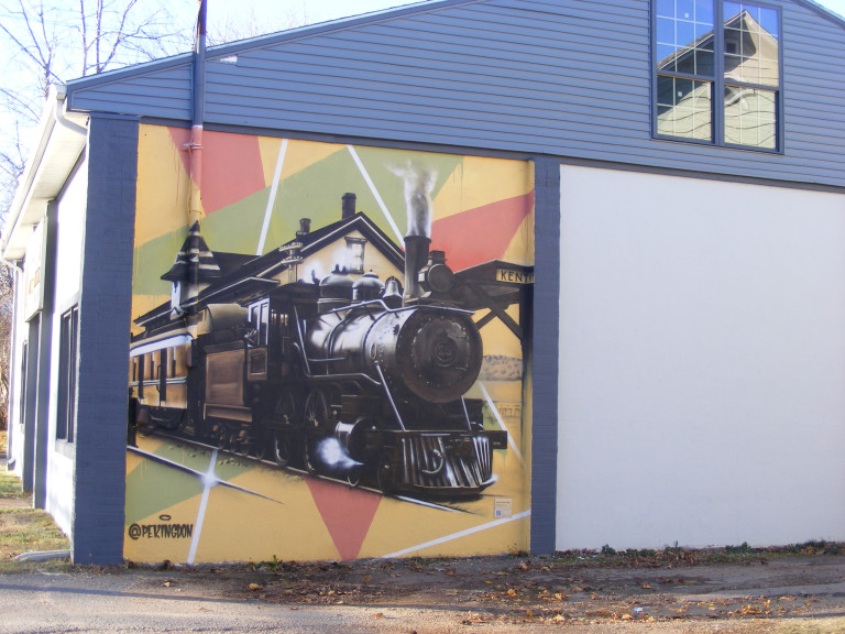 A black steam engine, with a train station in the background, is painted onto a green and yellow triangular background.