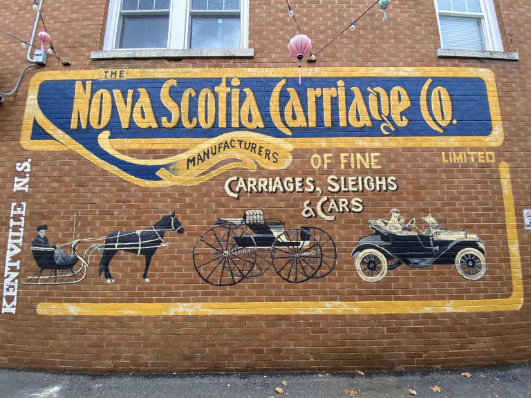 The side of a building is painted with images of a horse drawn sleigh, carriages and cars. "The Nova Scotia Carriage Co -  Manufacturers of Fine Carriages, Sleighs and Cars" is written at the top in old timey font.