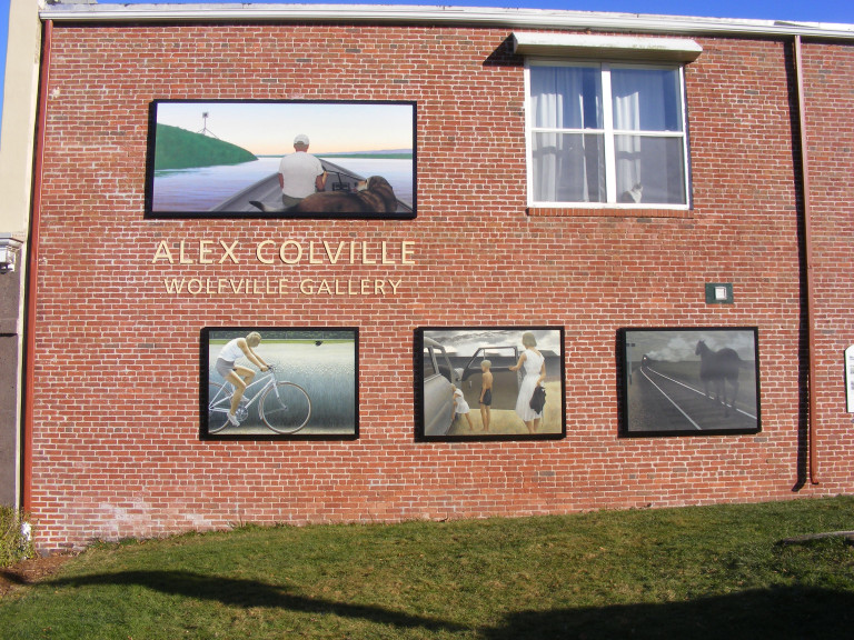 Paintings dot a red brick wall. Gold letters read "Alex Colville Wolfville Gallery." The paintings depict a man and his dog on a boat, a man on his bike, a woman and child getting into an open car door, and a black horse on train tracks.